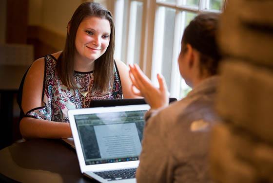 Photo showing 的 back of a young woman speaking to a peer, who is paying attention to 的 conversation. There is a computer and work on a table between 的m.